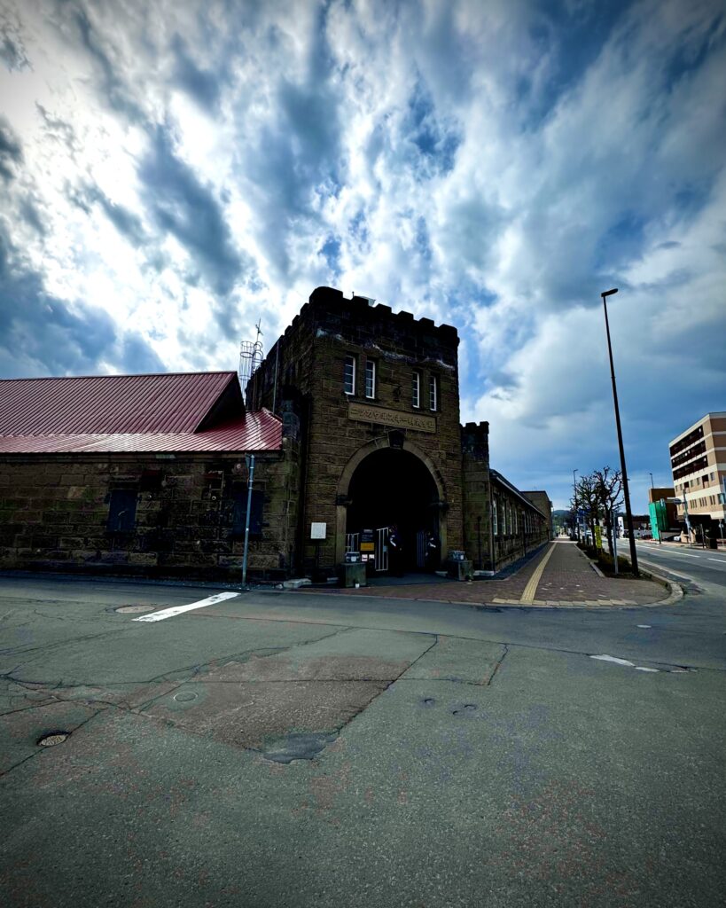 Stone building with arched entrance and red metal roof under a cloudy blue sky on a quiet street corner