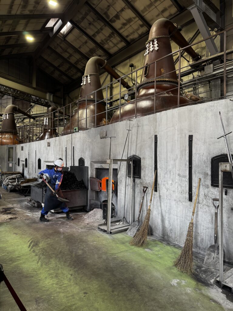 Worker shoveling coal into a glowing furnace below large copper distillation stills in an industrial building