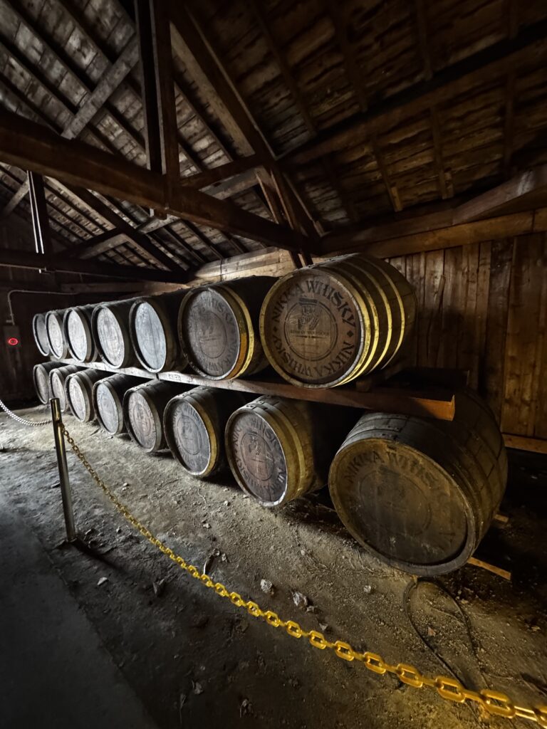 Stacked whisky barrels inside a wooden storage room with a chain barrier in front and slanted rafters above
