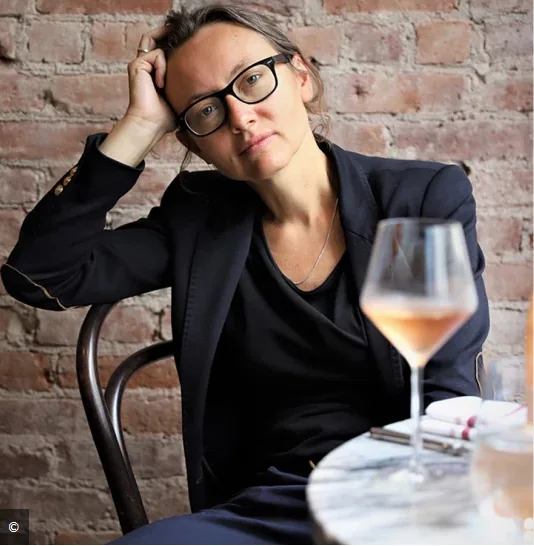 Person with glasses in black clothing seated at a table with a wine glass and papers, posing with hand on head