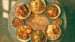 Overhead photograph of a round weathered wooden table with small shared Thai dishes, including grilled pork skewers, green papaya salad, chili relish, peanuts and rice, arranged in a ring around an empty center