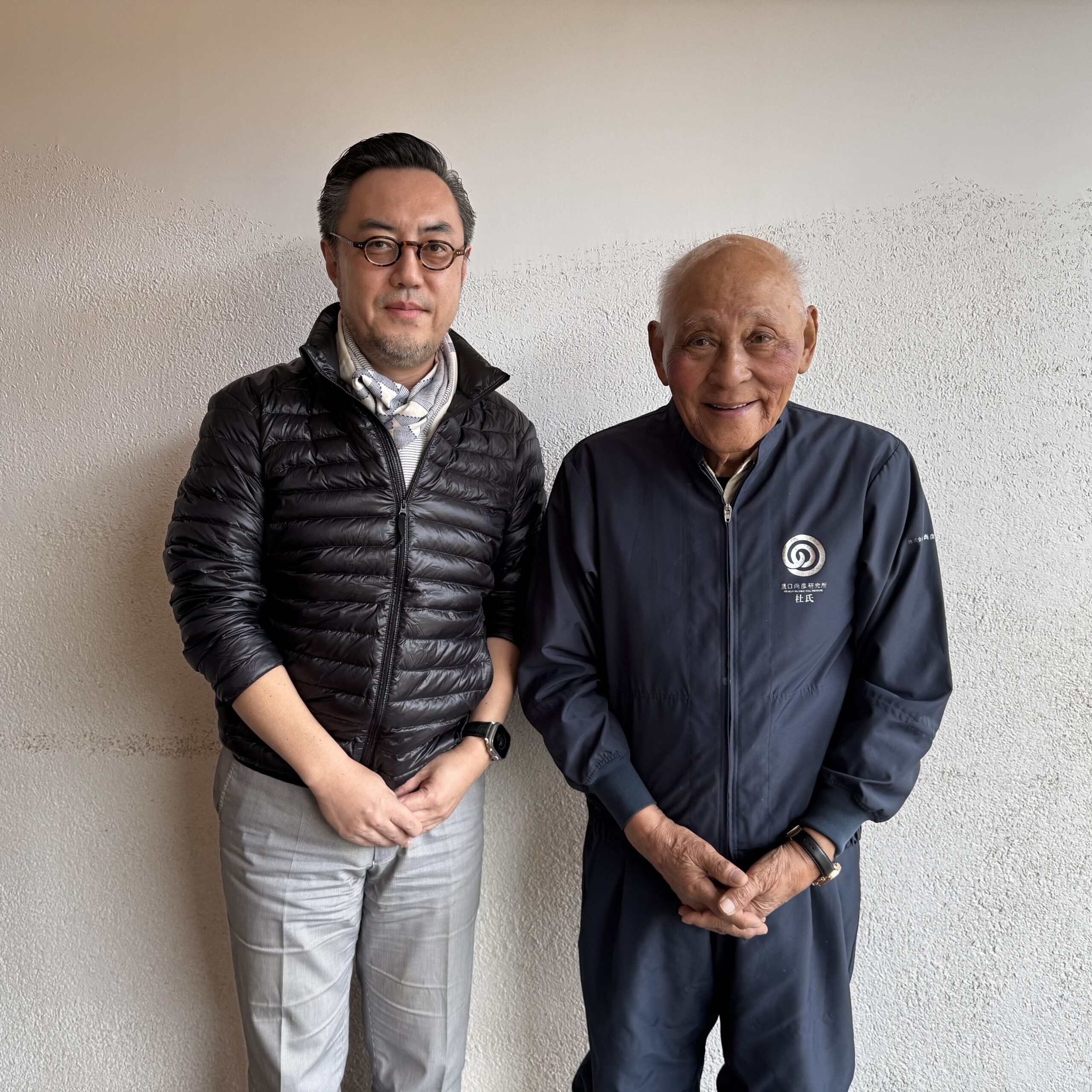 白い壁の前に並んで立つ二人。右は農口尚彦研究所の作業着を着た農口尚彦氏
